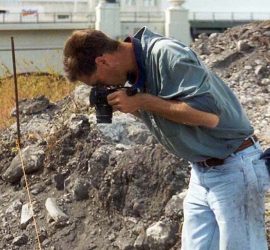 Image of the author shooting pictures at the site. He is wearing dirty blue jeans and a green work shirt. He has closely cropped hair. The Brickell Bridge is in the background and piles of rock and rubble are present.